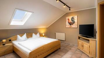 A bedroom with a sloped ceiling, a skylight, a bed with white bedding, a wooden headboard, bedside lamps, a radiator, a television on a wooden cabinet, and a rooster painting on the wall.
