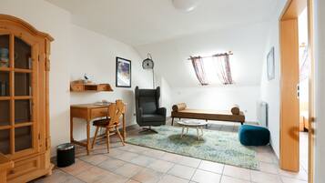 Una habitación con un armario de madera, un escritorio, una silla, un sillón negro, una mesa redonda de centro, un reposapiés azul, una alfombra estampada, un radiador y una ventana con cortinas.