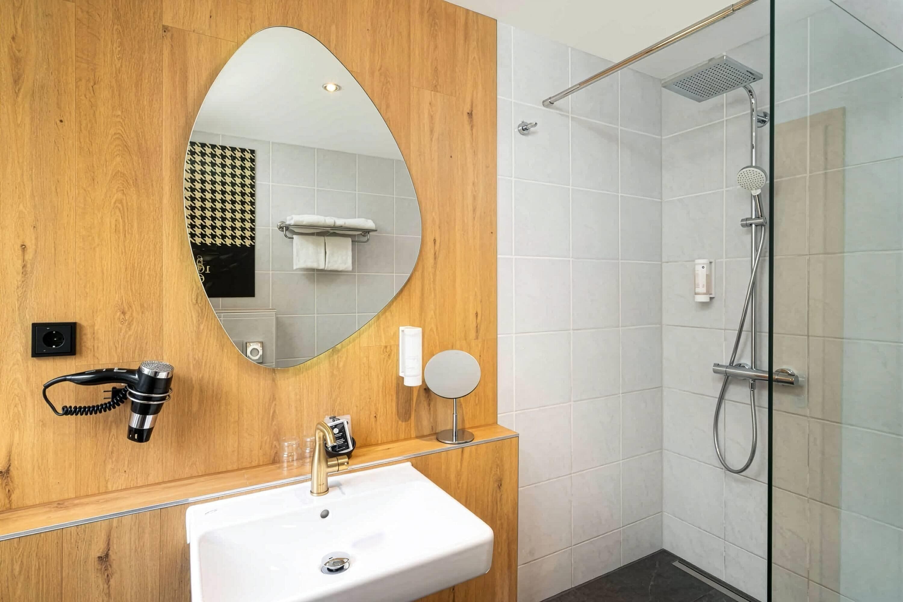 A modern bathroom with a wooden vanity, a large mirror, a handheld hair dryer, a sink, a glass shower enclosure, and a towel rack.