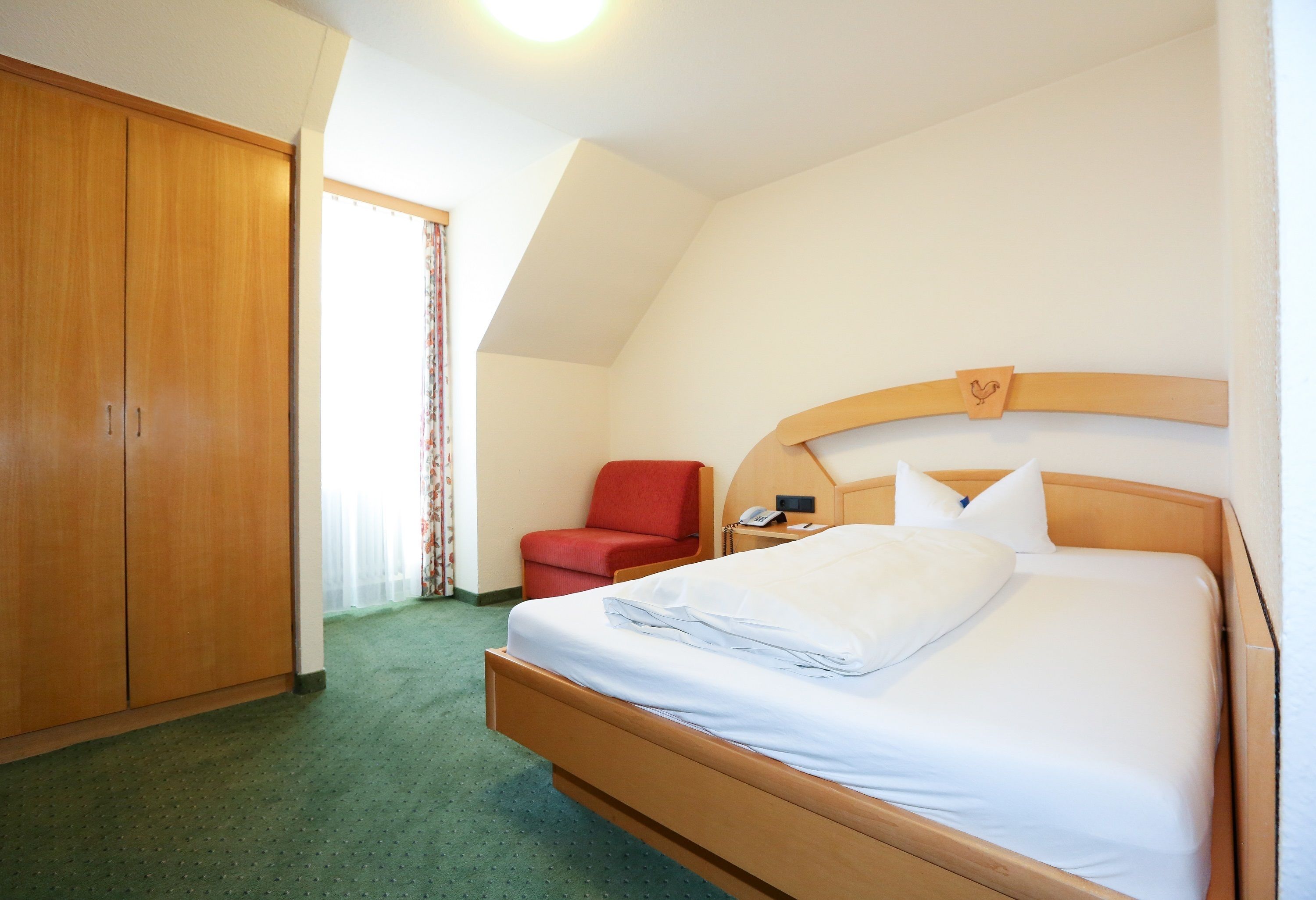 A hotel room with a wooden bed, a red chair, a wardrobe, and a telephone on the nightstand.
