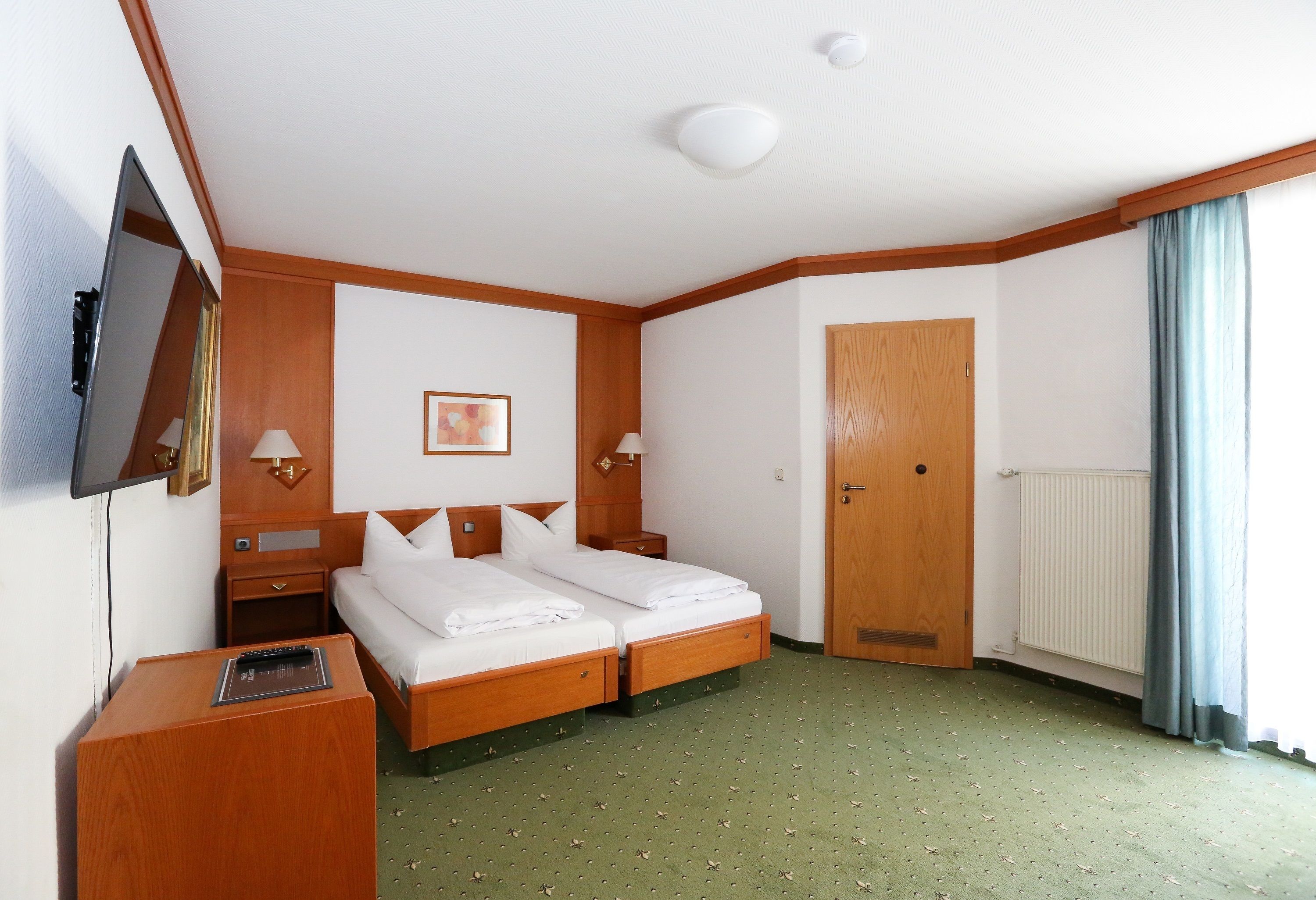 A hotel room with a double bed, a TV mounted on the wall, a desk with a lamp, and a wooden door.