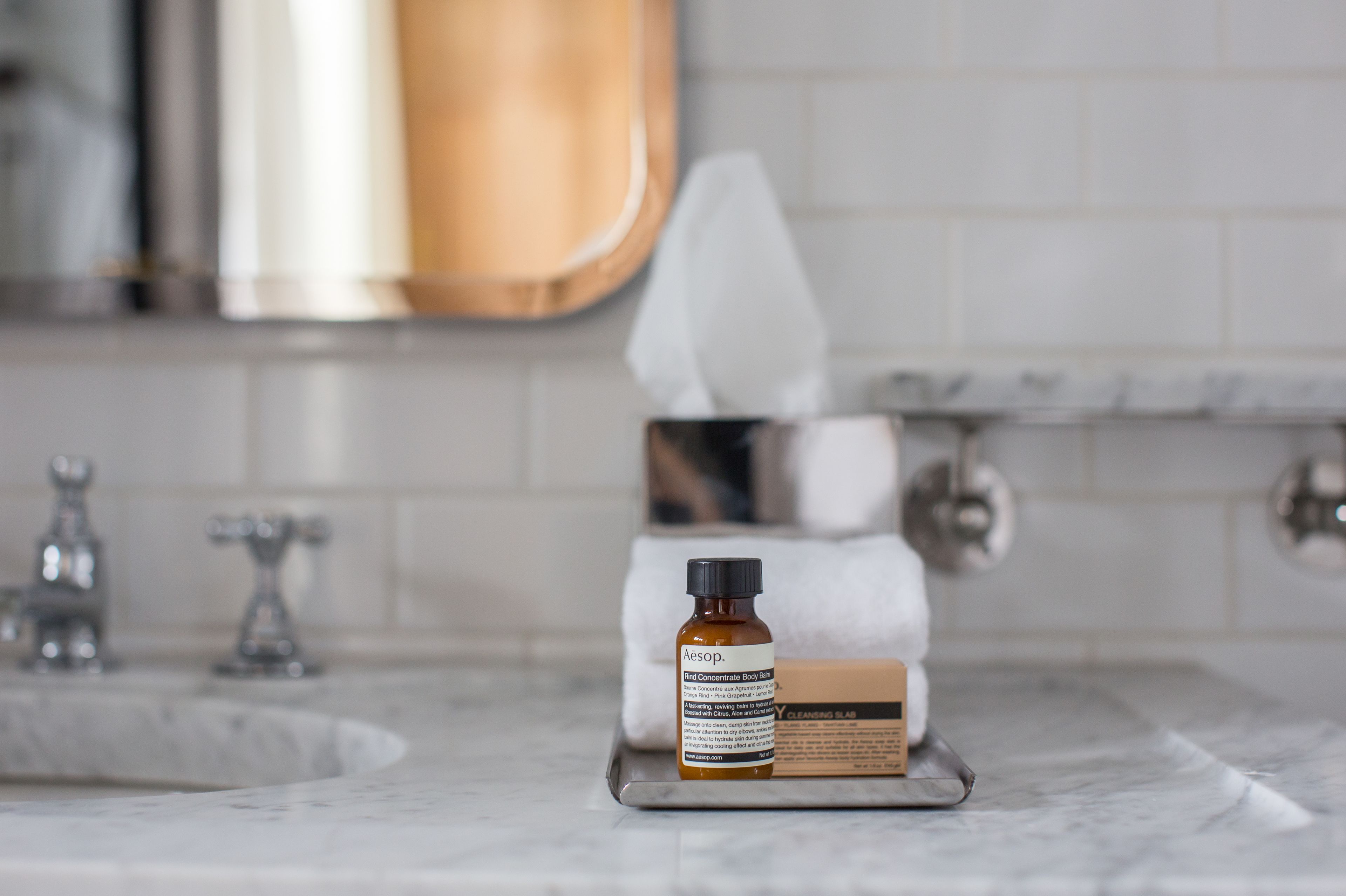 A bathroom with a marble sink, a bottle of body serum, and a box of body wash on a tray.