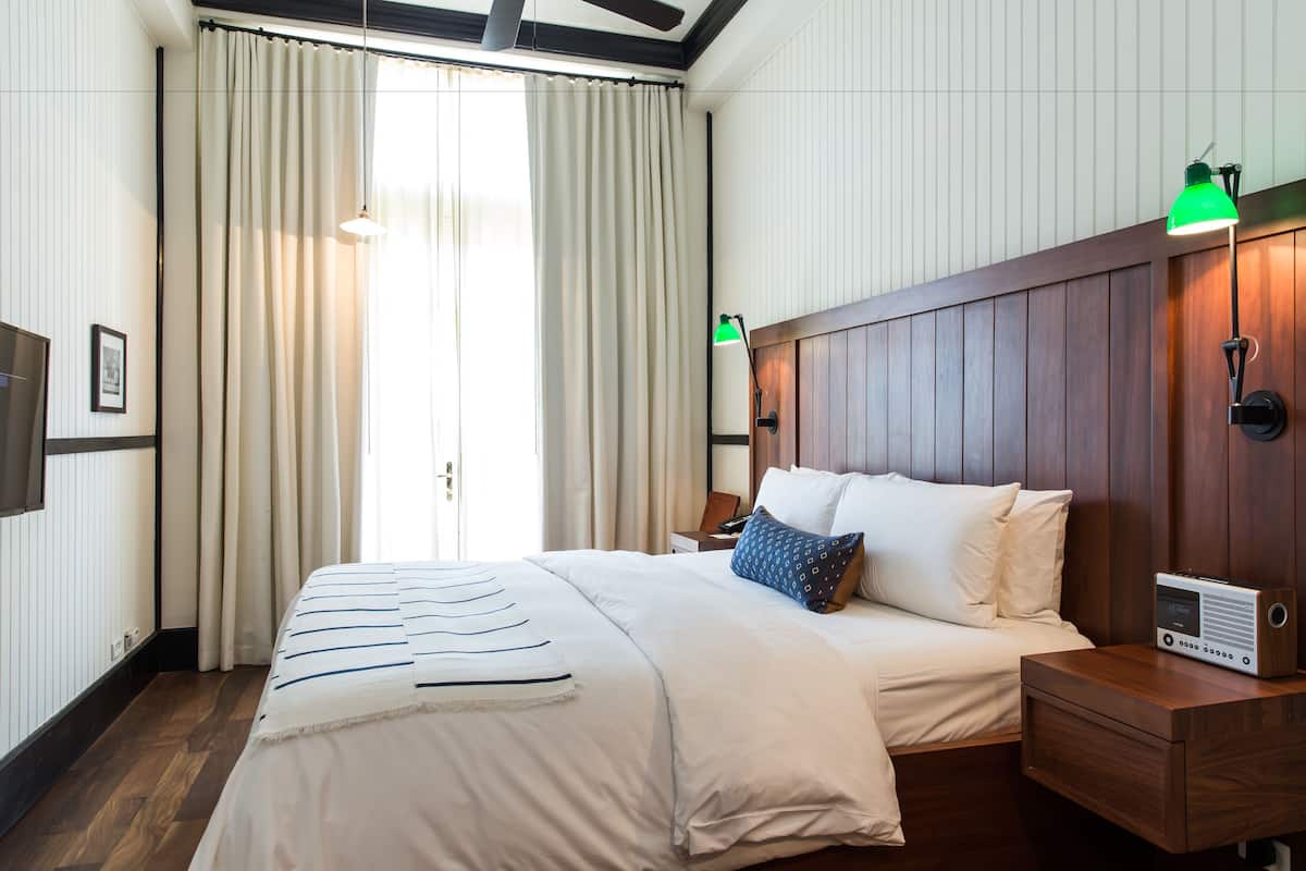 A hotel room with a large bed, wooden headboard, a bedside table with a radio, and a window with curtains.
