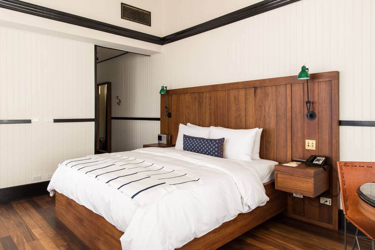 A hotel room with a wooden headboard, a bed with white bedding, a nightstand with a lamp, and a chair.