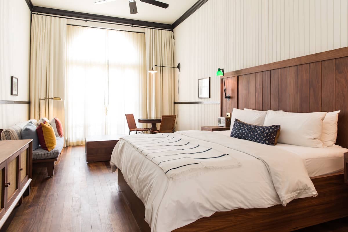 A hotel room with a large bed, a wooden headboard, a desk with a chair, a sofa with cushions, and a large window with curtains.
