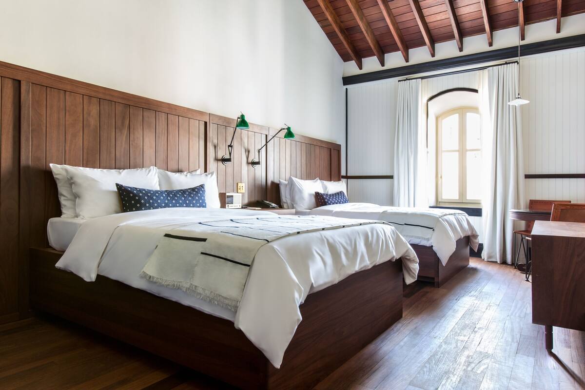 A double bed with white linens and two pillows, a wooden headboard, a nightstand with a lamp, and a window with curtains.