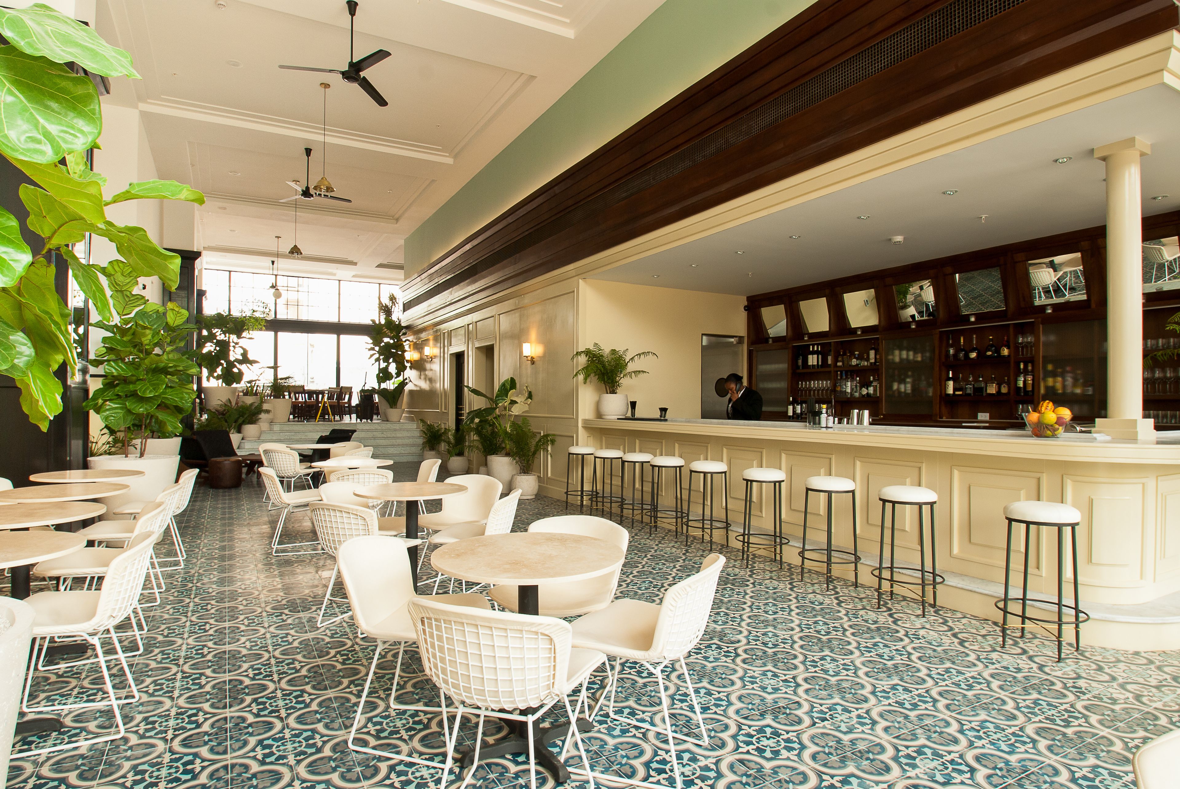 Zona de bar con taburetes altos y barra, rodeada de sillas y mesas blancas, piso estampado y plantas grandes en macetas.