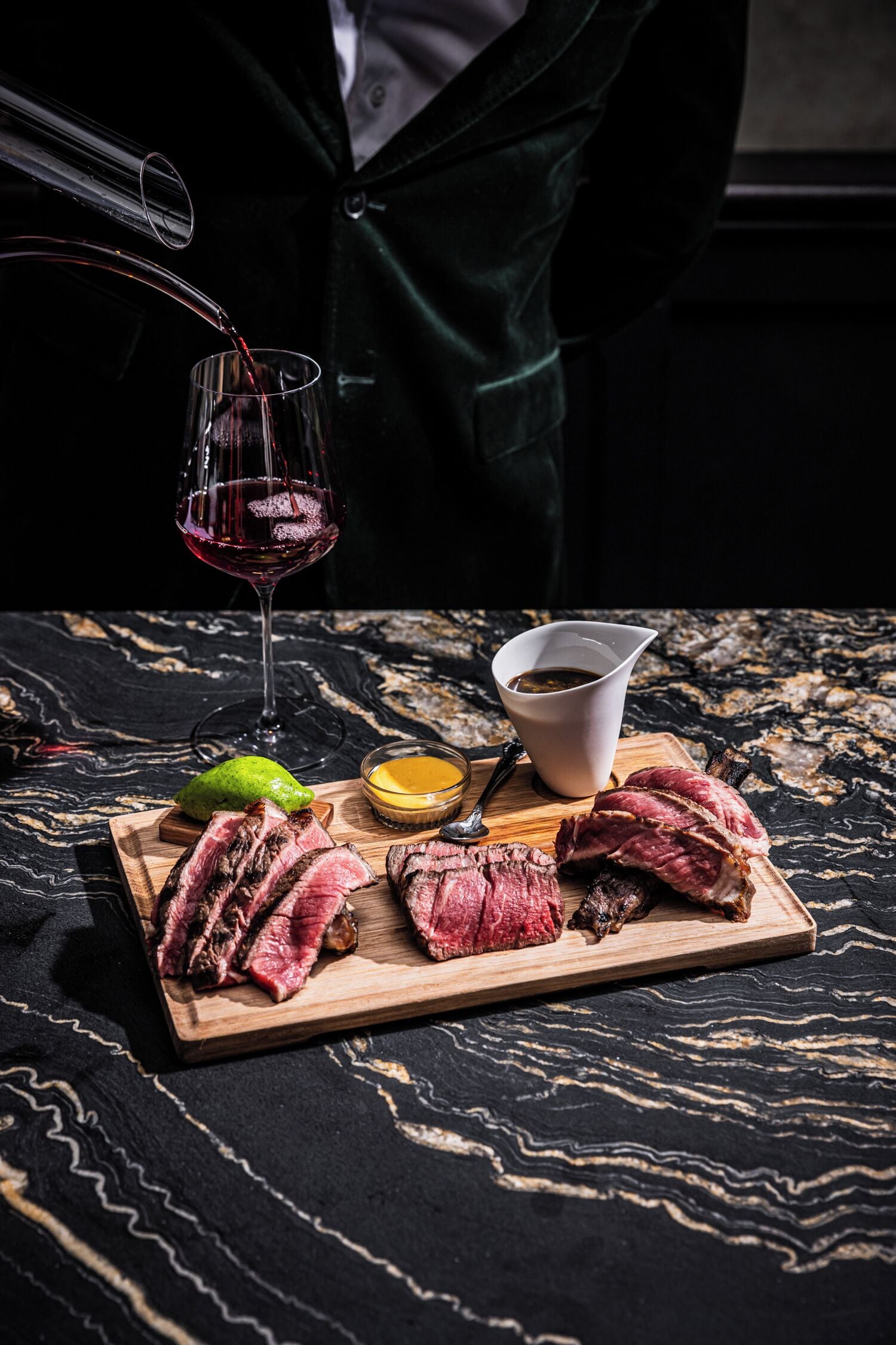 Una tabla de madera con bistec en rodajas, una copa de vino tinto y salsas en una mesa de mármol.