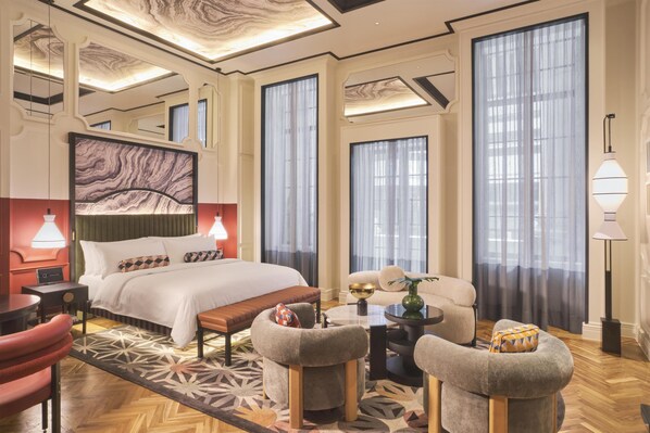 Una habitación de hotel moderna con una cama grande, una zona de estar con un sofá y sillones, una mesa de centro y un ventanal con cortinas.