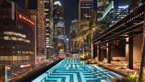 Vista nocturna de una zona urbana moderna con una larga piscina iluminada en primer plano, rodeada de edificios altos y palmeras.