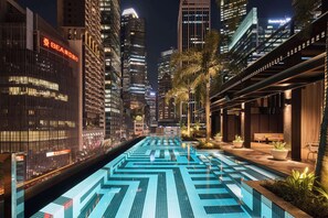 Vista nocturna de una zona urbana moderna con una larga piscina iluminada en primer plano, rodeada de edificios altos y palmeras.