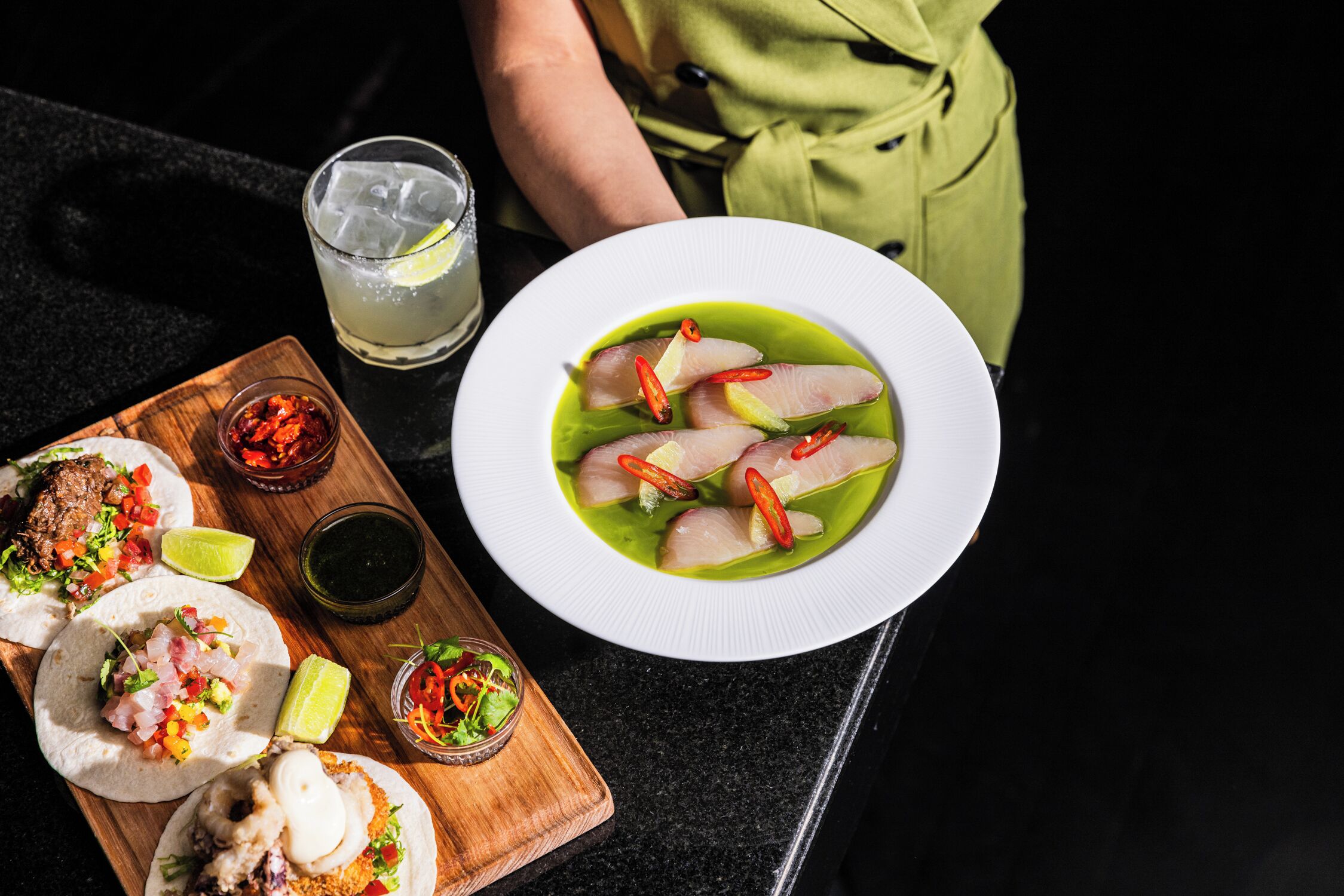 Una mesa con un plato de sopa verde, un vaso de bebida helada y varios platillos, entre ellos tacos y un tazón pequeño de salsa.