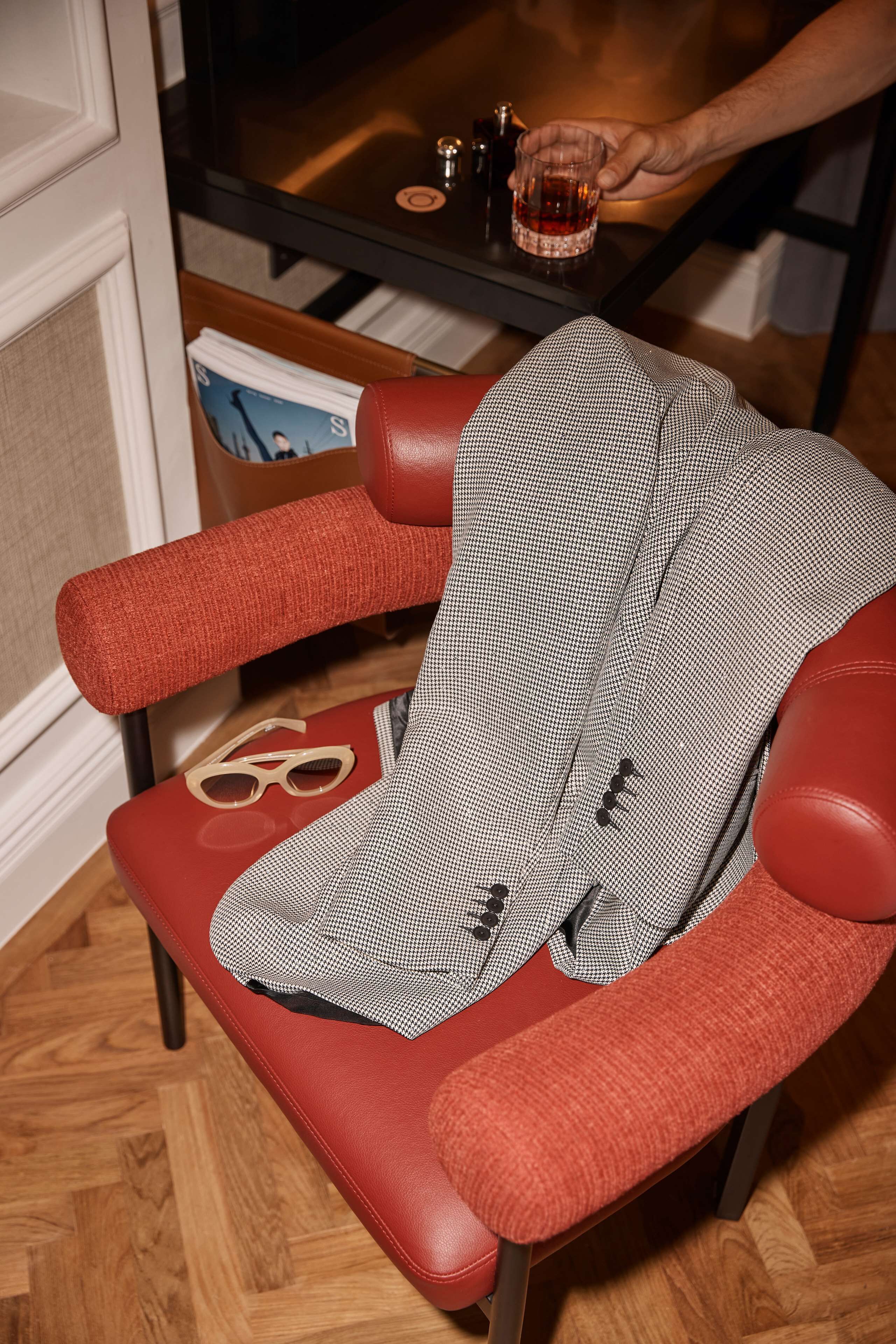 Une chaise rouge, recouverte d’une veste à carreaux, une paire de lunettes et un verre de whisky posés sur une table.
