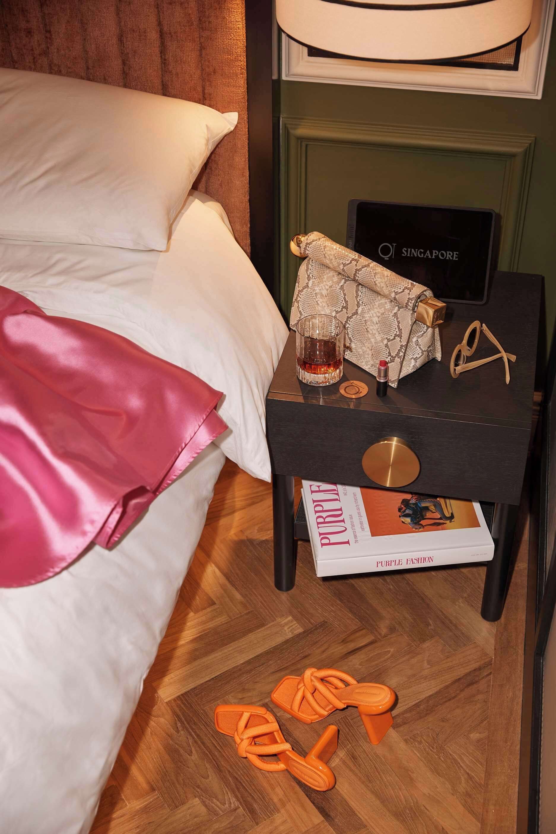 A neatly made bed with white linens, a small bedside table with a magazine, a handbag, and a pair of slippers.