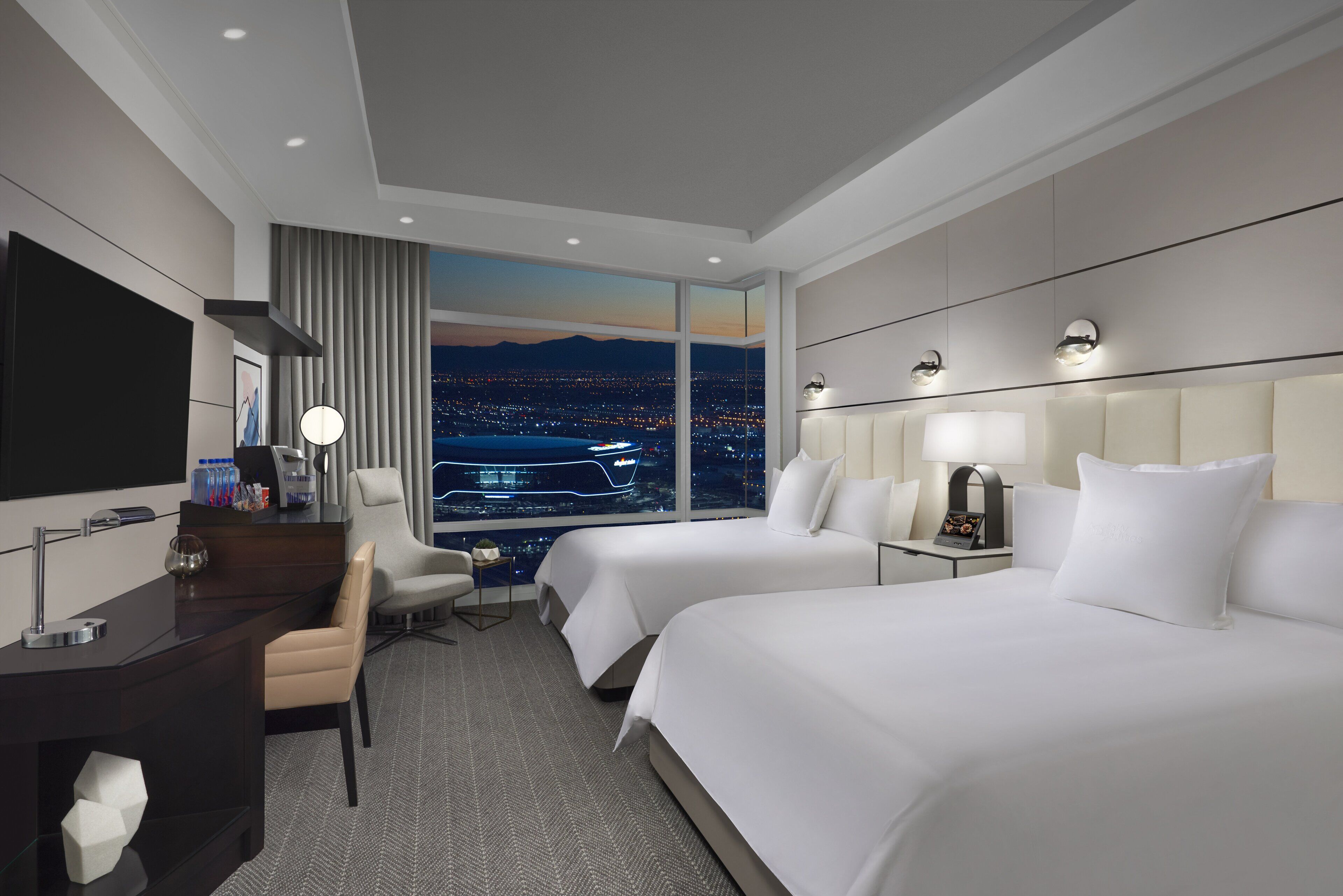 Ein Hotelzimmer mit zwei Betten, einem Schreibtisch, einem Stuhl, einem Fernseher und einem großen Fenster mit Blick auf die Stadt.