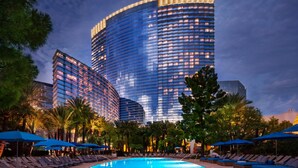 A modern hotel with a curved glass facade, a pool area with lounge chairs and umbrellas, and palm trees. - ARIA Sky Suites (Las Vegas)