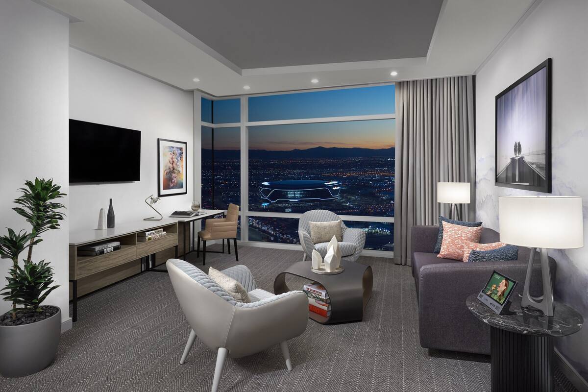 Una habitación de hotel moderna con un amplio ventanal que ofrece vistas a la ciudad al atardecer, un televisor de pantalla plana, un escritorio con lámpara y asientos cómodos con una mesita de centro.