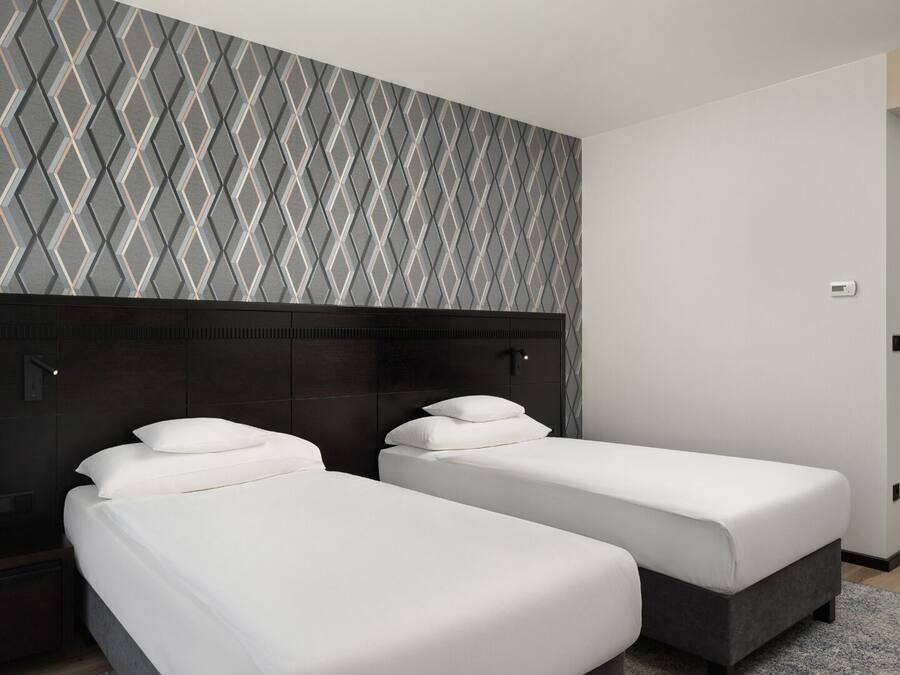 A hotel room with two single beds, a dark headboard, a geometric patterned wall, and a door to the right.