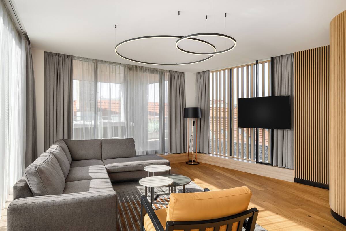 Ein modernes Wohnzimmer mit einer Eckcouch, zwei kleinen runden Tischen, einem Flachbildfernseher und großen Fenstern mit Vorhängen.