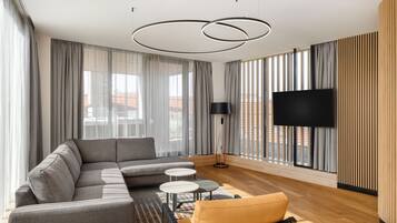 Una sala de estar moderna con un sofá modular, dos mesitas redondas, un televisor de pantalla plana y ventanas amplias con cortinas.