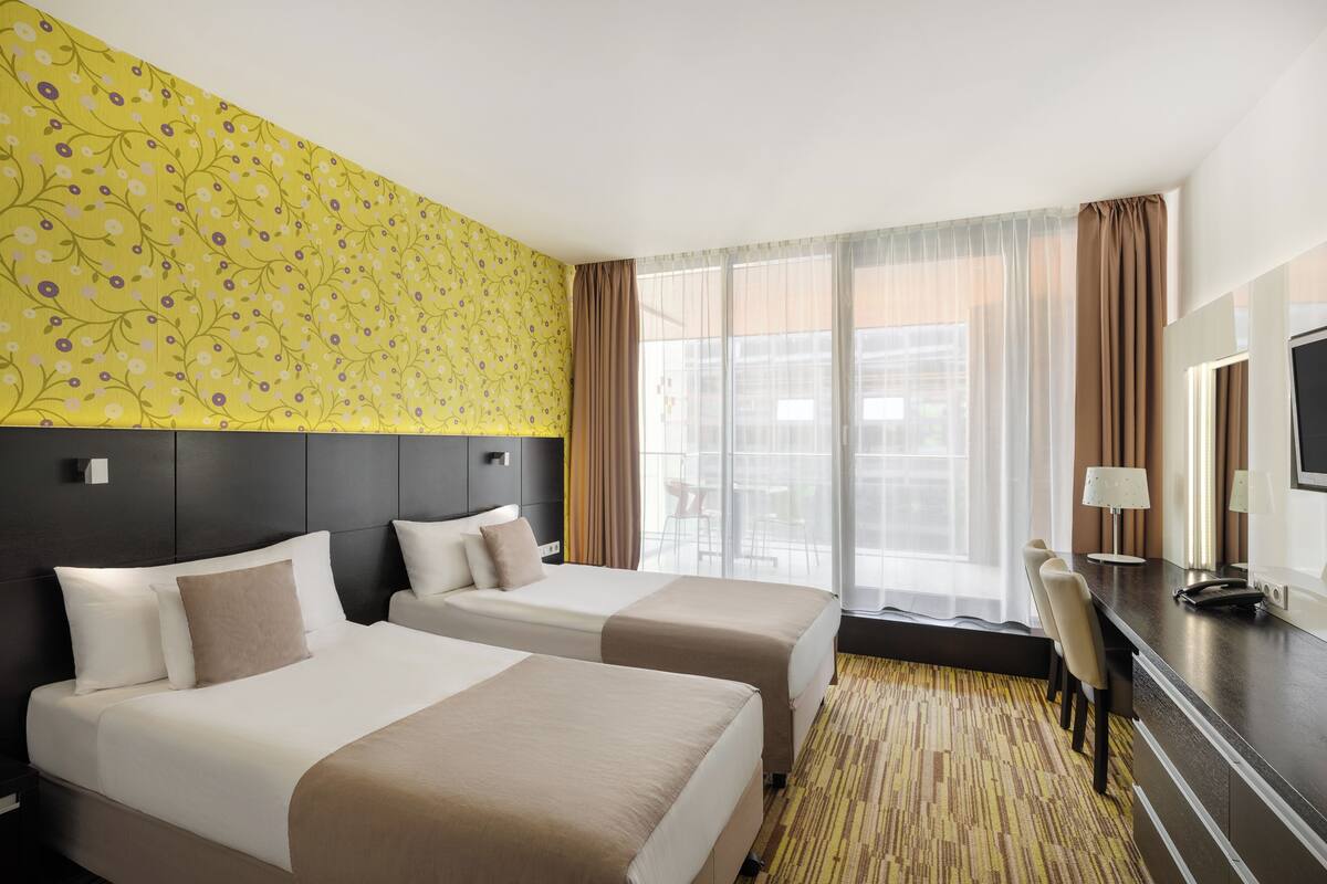 Ein Hotelzimmer mit zwei Betten, einem Schreibtisch und einem großen Fenster mit Vorhängen.