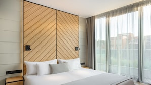 Una habitación de hotel moderna con una cama grande, cabecera de madera, mesitas de noche y vistas a través de cortinas translúcidas.