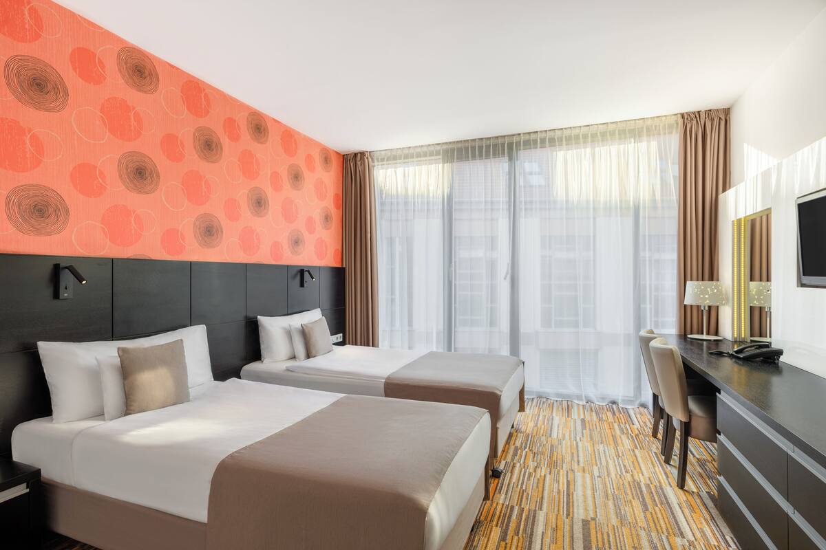 Ein Hotelzimmer mit zwei Betten, einem Schreibtisch, einem Sessel, einem Fernseher und einem großen Fenster mit Vorhängen.