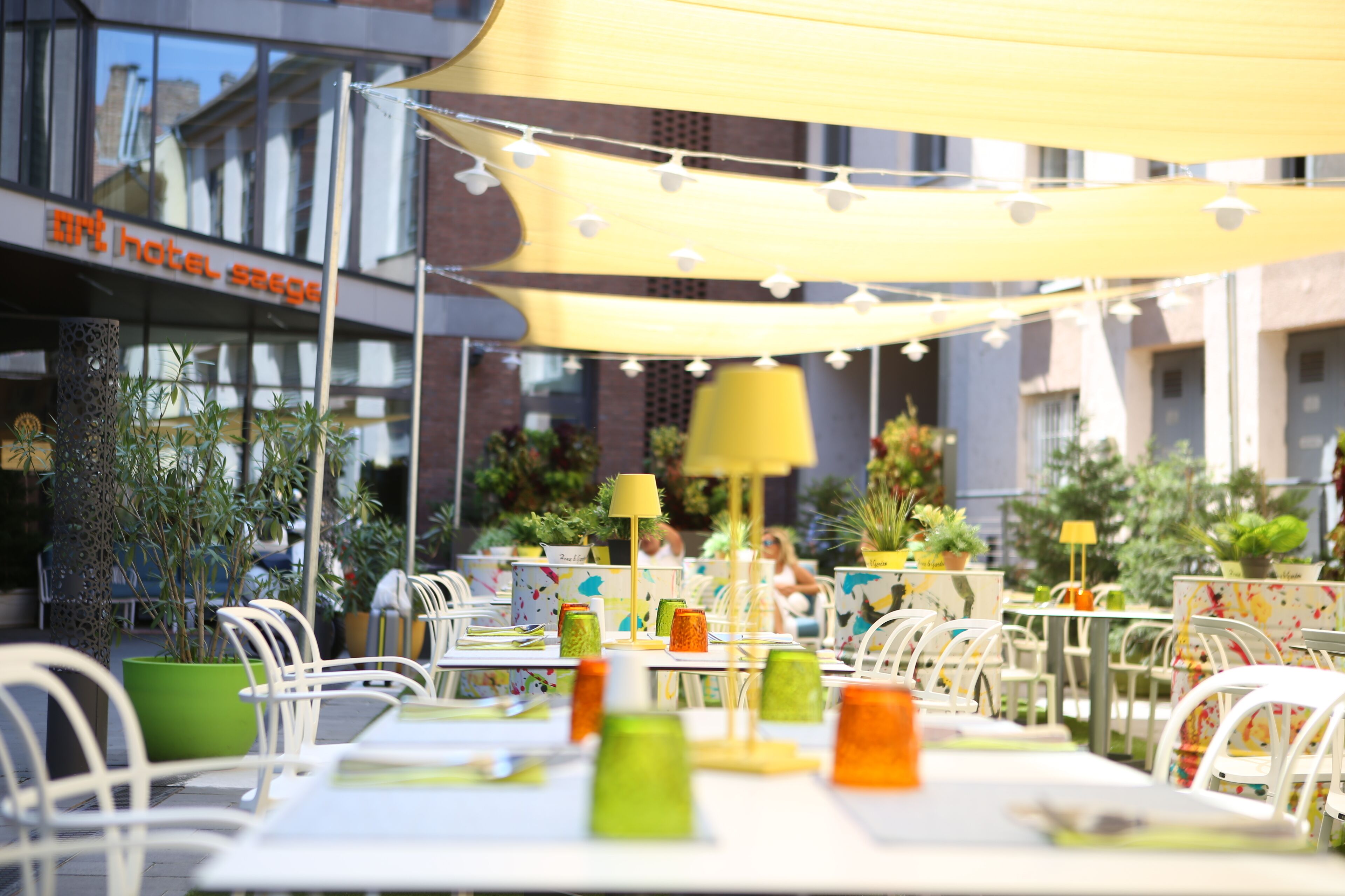 An outdoor cafe with tables and chairs, string lights, and potted plants.