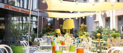 An outdoor cafe with tables and chairs, string lights, and potted plants.