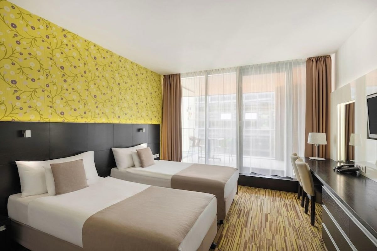 Una habitación de hotel con dos camas, un escritorio y un ventanal con cortinas.