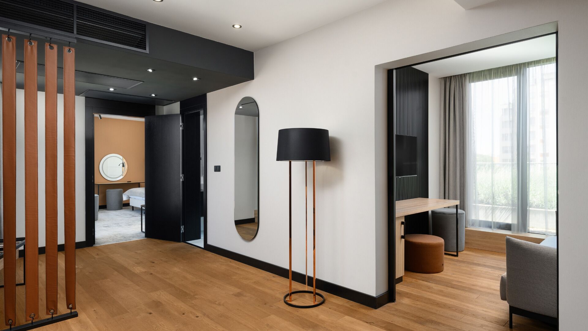 Una habitación de hotel moderna con piso de madera, un espejo grande, una lámpara de pie, un televisor y un área de descanso con vistas al exterior.
