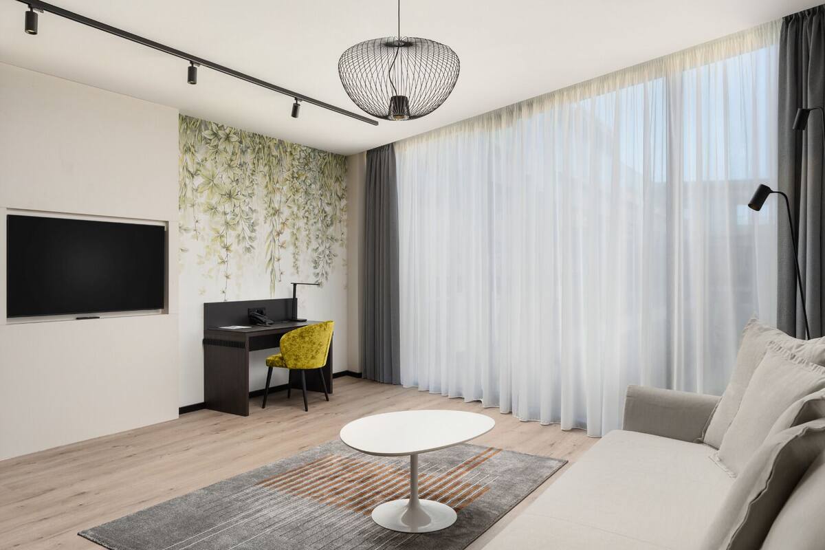 Ein modernes Wohnzimmer mit einem Flachbildfernseher, einem Schreibtisch, einem gelben Sessel, einem runden weißen Tisch und einem großen Fenster mit durchsichtigen Vorhängen.