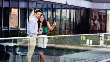 A couple standing on a balcony with a glass railing, overlooking a modern building with large windows and a reflection of a sculpture.