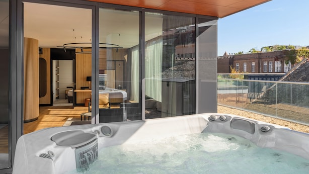 Un jacuzzi moderno en el interior, con recinto de vidrio, techo de madera y vista a un edificio con tejado inclinado.
