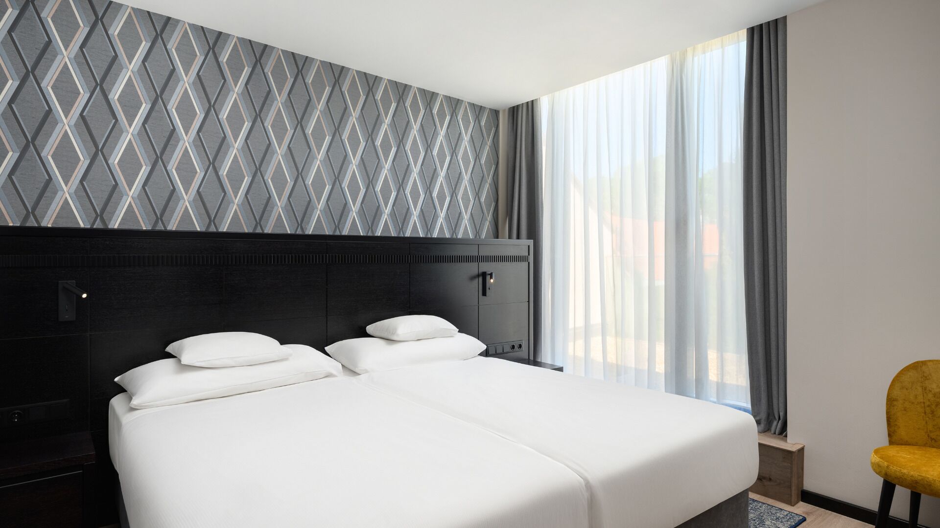 A modern hotel room with a large bed, a headboard with a geometric pattern, a bedside table, and a view of the outdoors through sheer curtains.