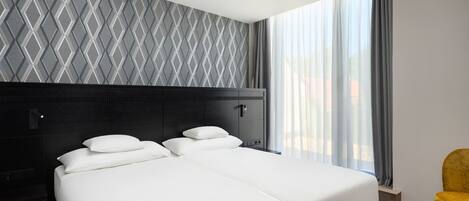 Una habitación de hotel moderna con una cama grande, cabecera con diseño geométrico, una mesita de noche y vistas al exterior a través de cortinas translúcidas.