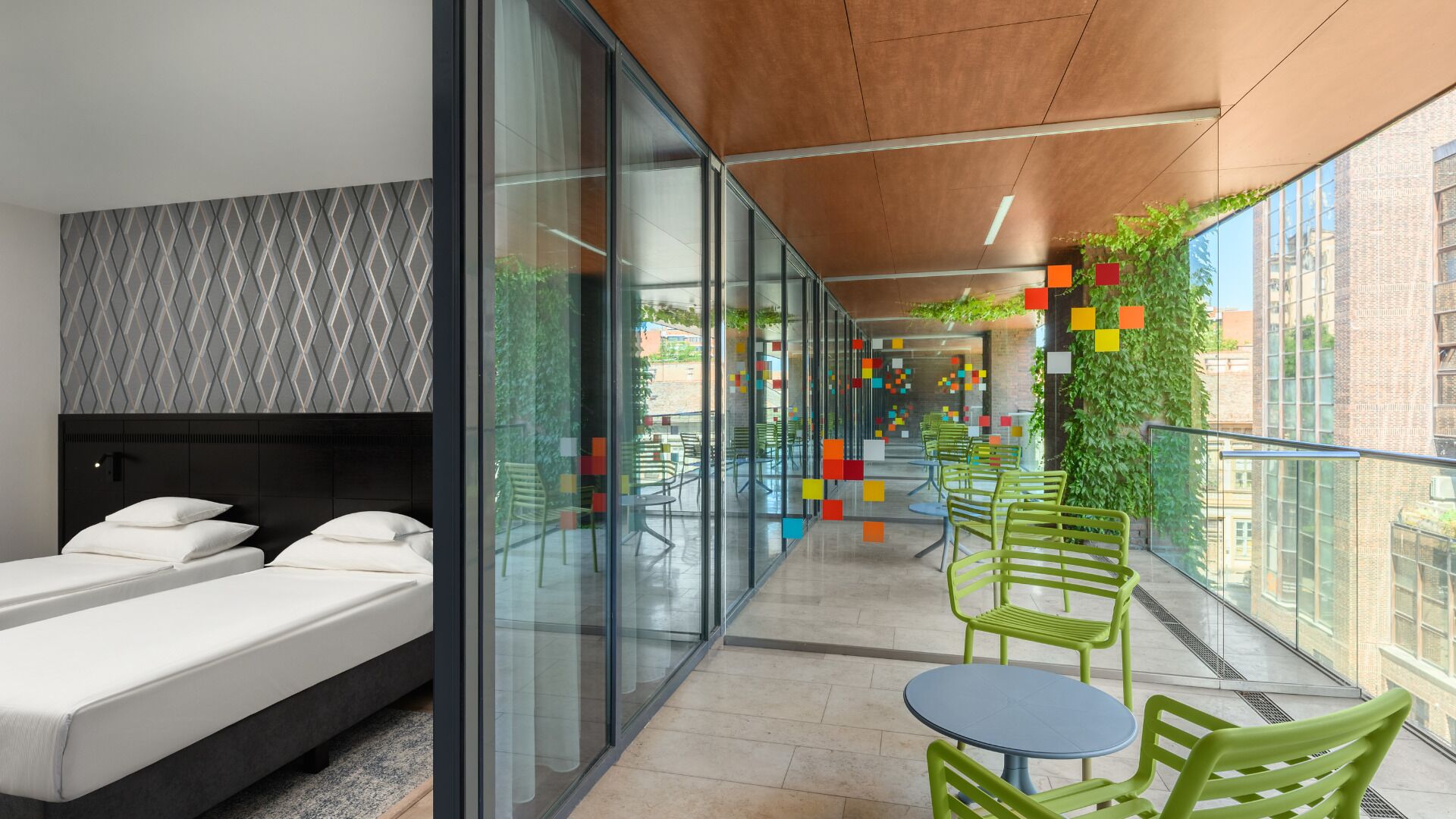 A modern hotel room with a bed, a glass wall, and a balcony with chairs and greenery.