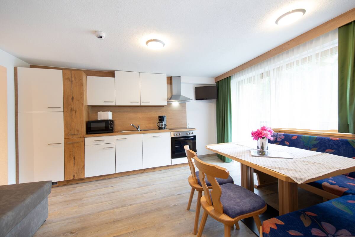 Una cocina compacta con armarios de madera, un microondas y una estufa. Hay una mesa de comedor con sillas y un banco con cojín estampado de flores.