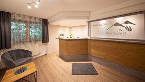 A reception area with a wooden reception desk, a grey sofa, and a small table with a plate. - Gästehaus Waldesrand (Soelden)