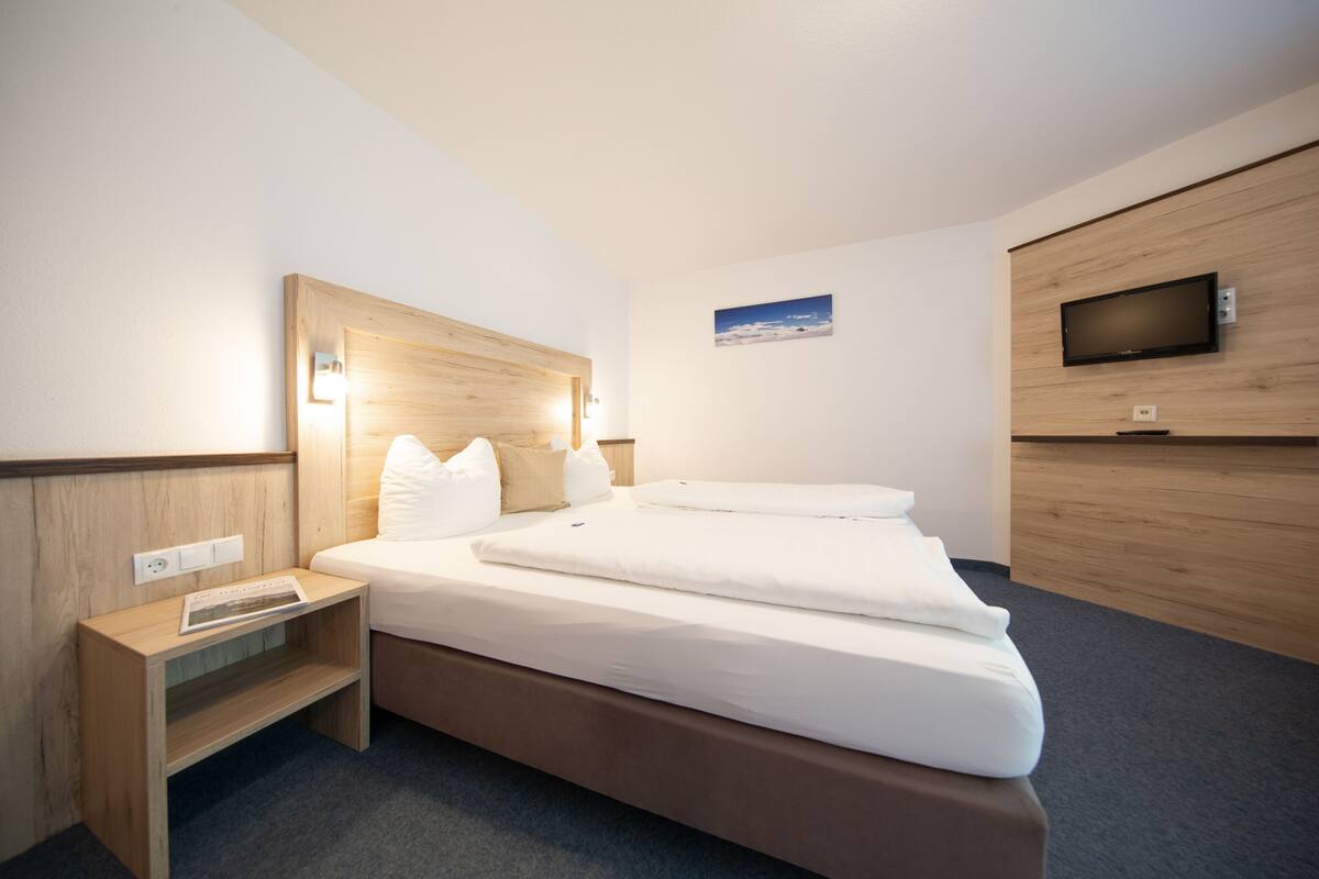 Una habitación de hotel moderna con un cabecero de madera, un televisor de pantalla plana montado en la pared, una cama bien hecha con sábanas blancas y una mesita de noche con una revista.