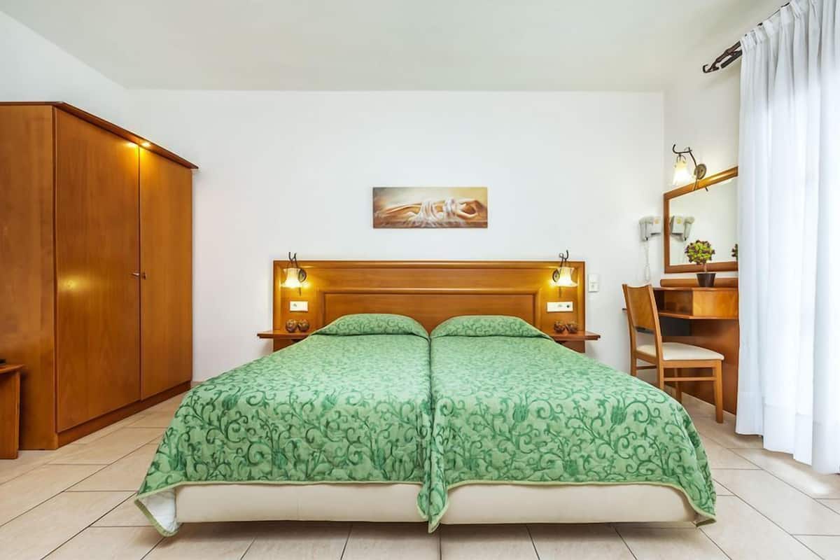 Un lit double avec une couvre-lit à motifs verts, une tête de lit en bois, des lampes de chevet, une armoire en bois, une petite table, une chaise, un miroir et un tableau accroché au mur.