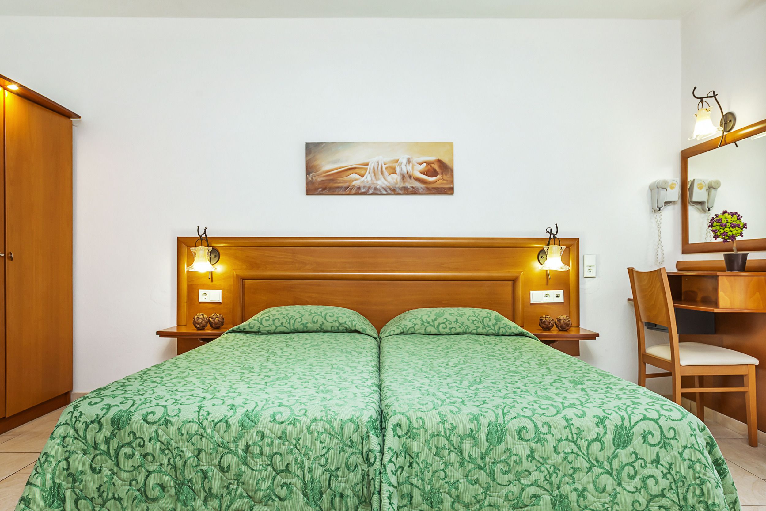 A room with a bed, two bedside tables with lamps, a wooden headboard, a painting on the wall, a chair, a desk, and a mirror.