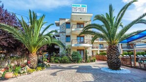 Un hôtel avec une enseigne indiquant « HÔTEL MHPHIDEZ » et une cour dallée agrémentée de palmiers et de plantes en pot.