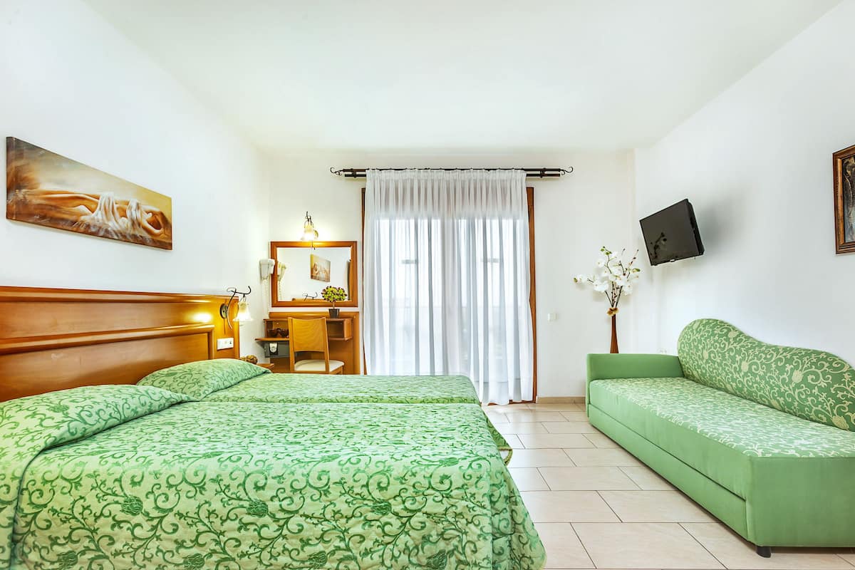 Une chambre d’hôtel avec deux lits, un canapé vert, un bureau avec une chaise, un téléviseur fixé au mur et une fenêtre avec des rideaux.