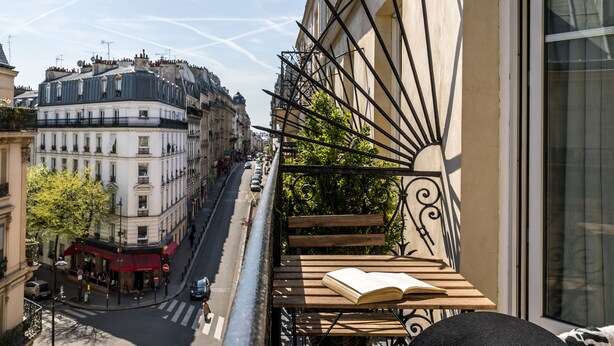 A balcony with a wooden table and an open book, overlooking a street with buildings and a café. - Hotel Boronali Montmartre (Paris)
