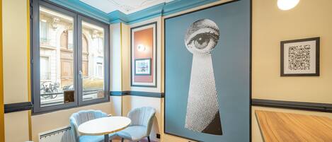 A room with a round table, two chairs, a large artwork on the wall, and a window with a view of the street.