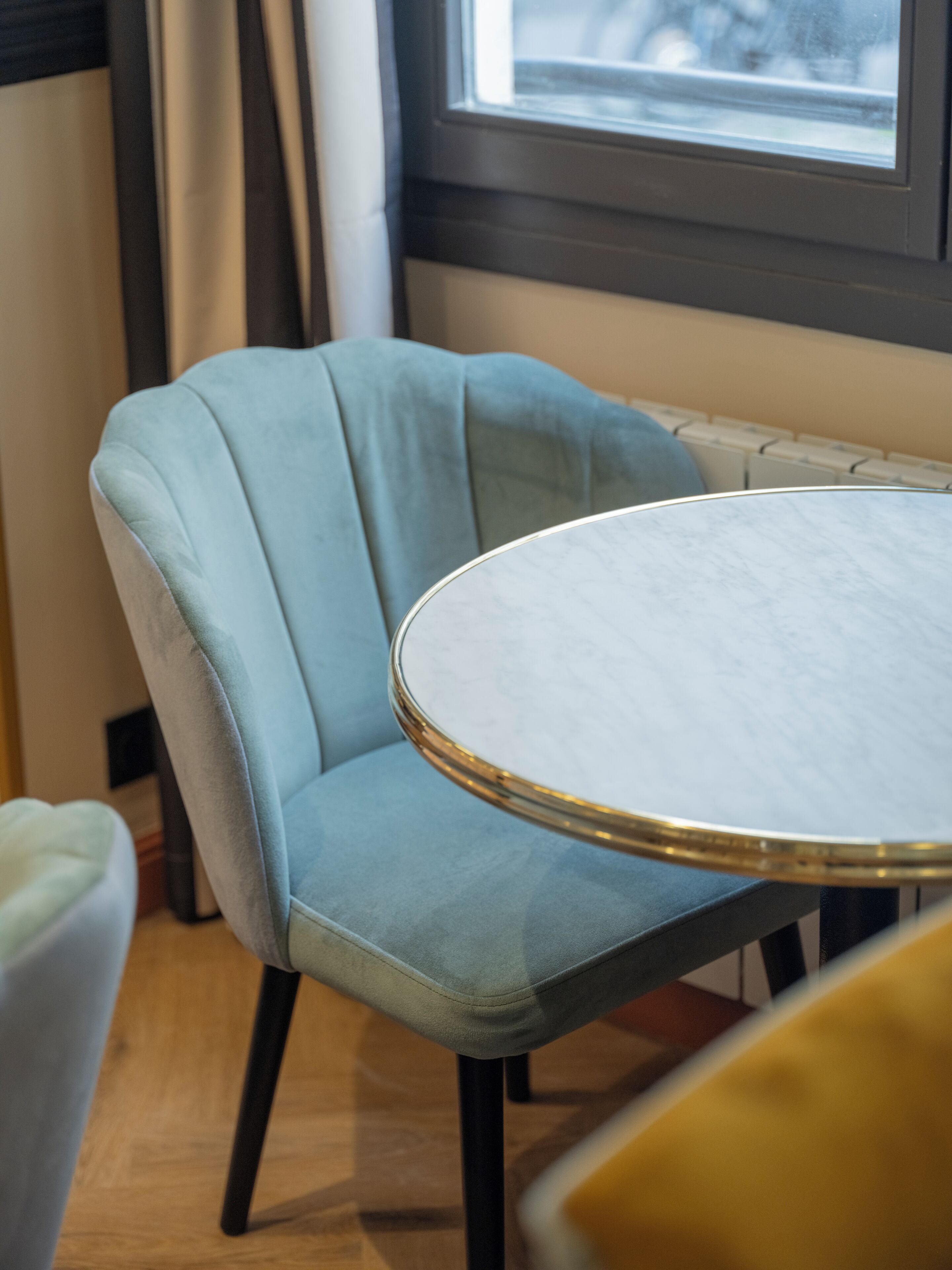 Une table ronde avec un plateau en marbre, une chaise bleu clair rembourrée avec un dossier incurvé, et un radiateur en arrière-plan.