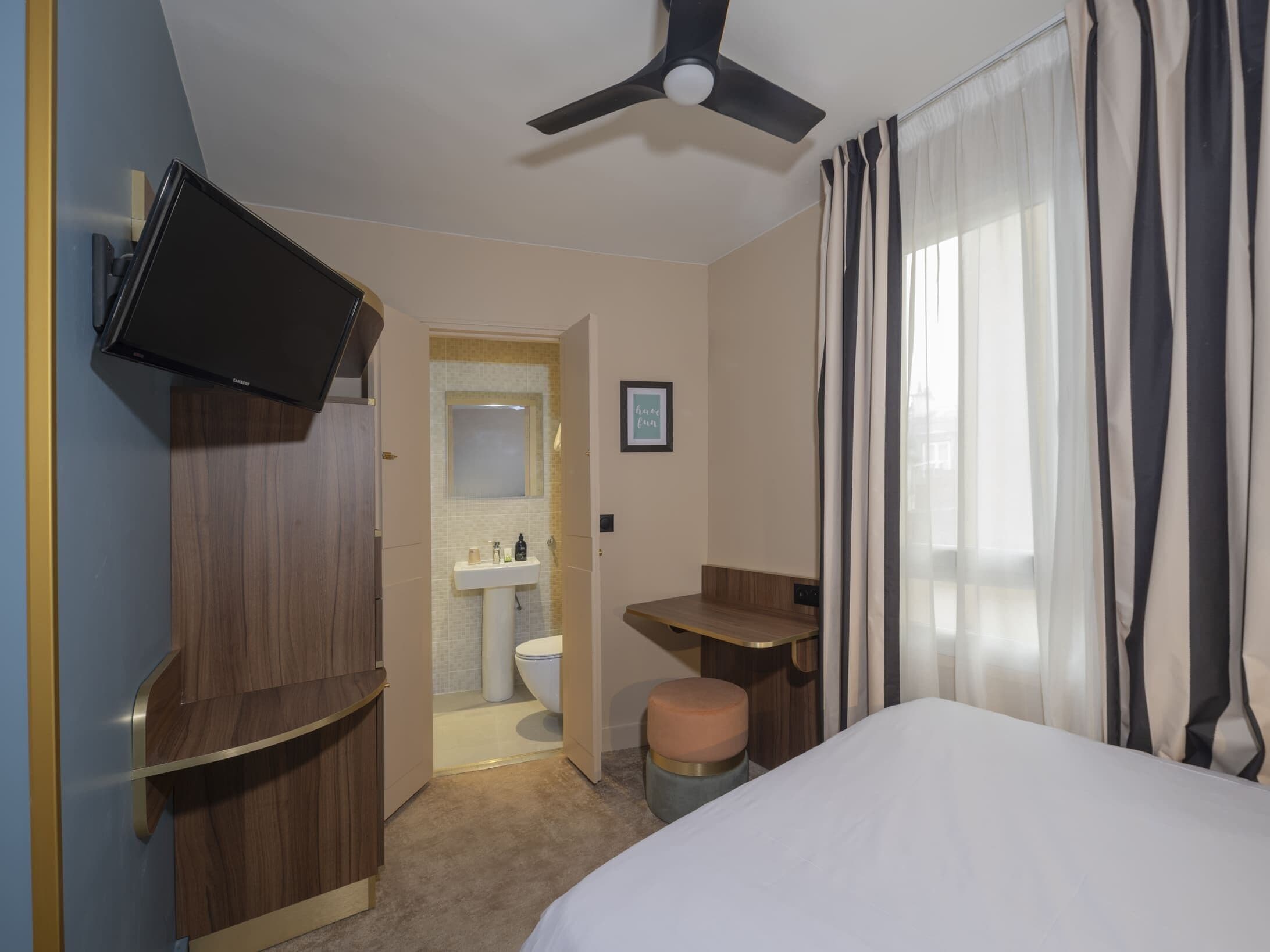 A hotel room with a bed, a television mounted on the wall, a wooden wardrobe, a small stool, and a bathroom visible through an open door.