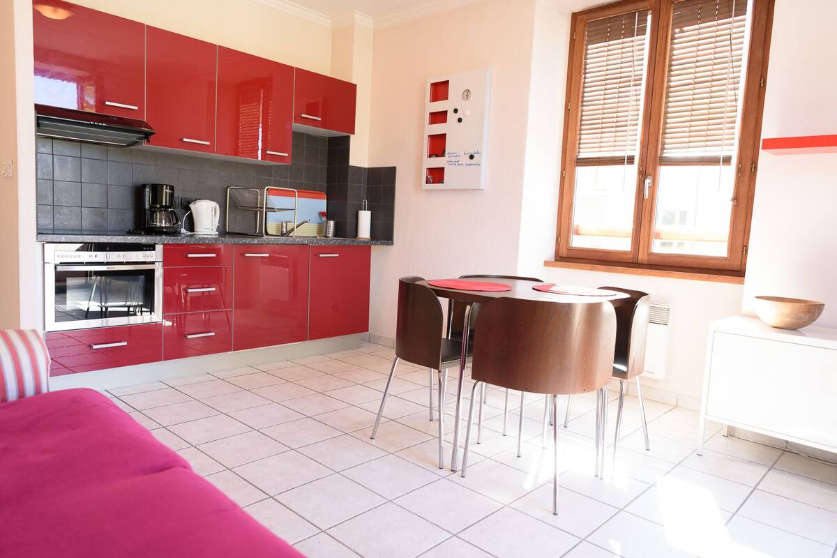 Una cocina moderna con armarios rojos, un comedor con mesa y sillas, y un ventanal con persianas.