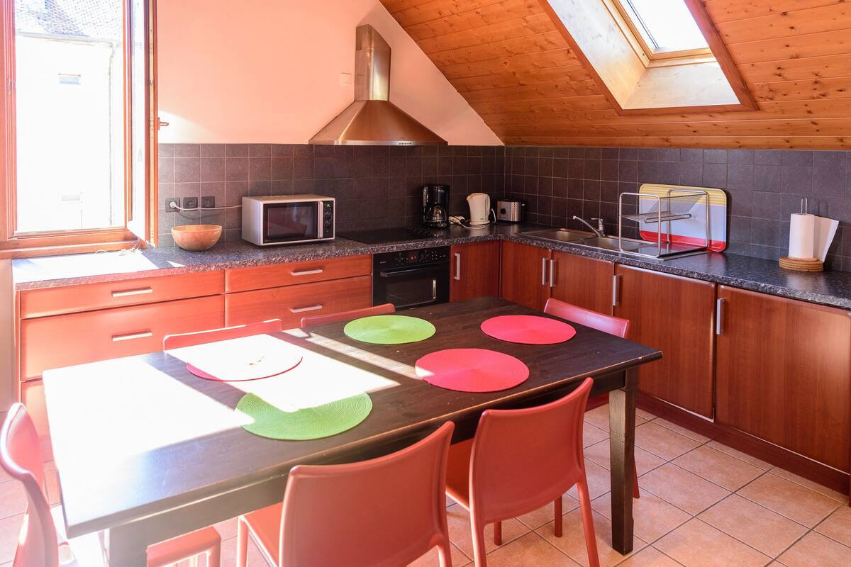 Una cocina con techo de madera, tragaluz, microondas, cafetera y una mesa de comedor con individuales de colores.